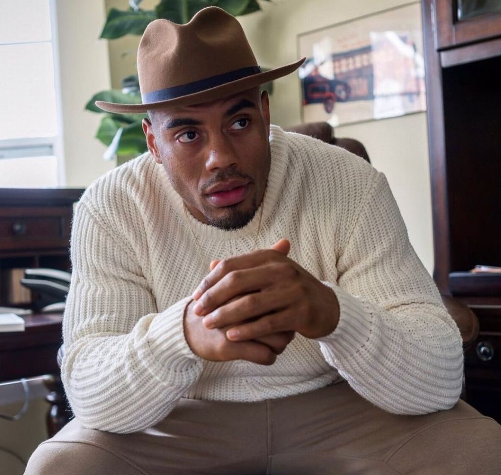 Rashad is wearing a hat, brown pants and white sweater in this picture. We can see a serious expression in his face.