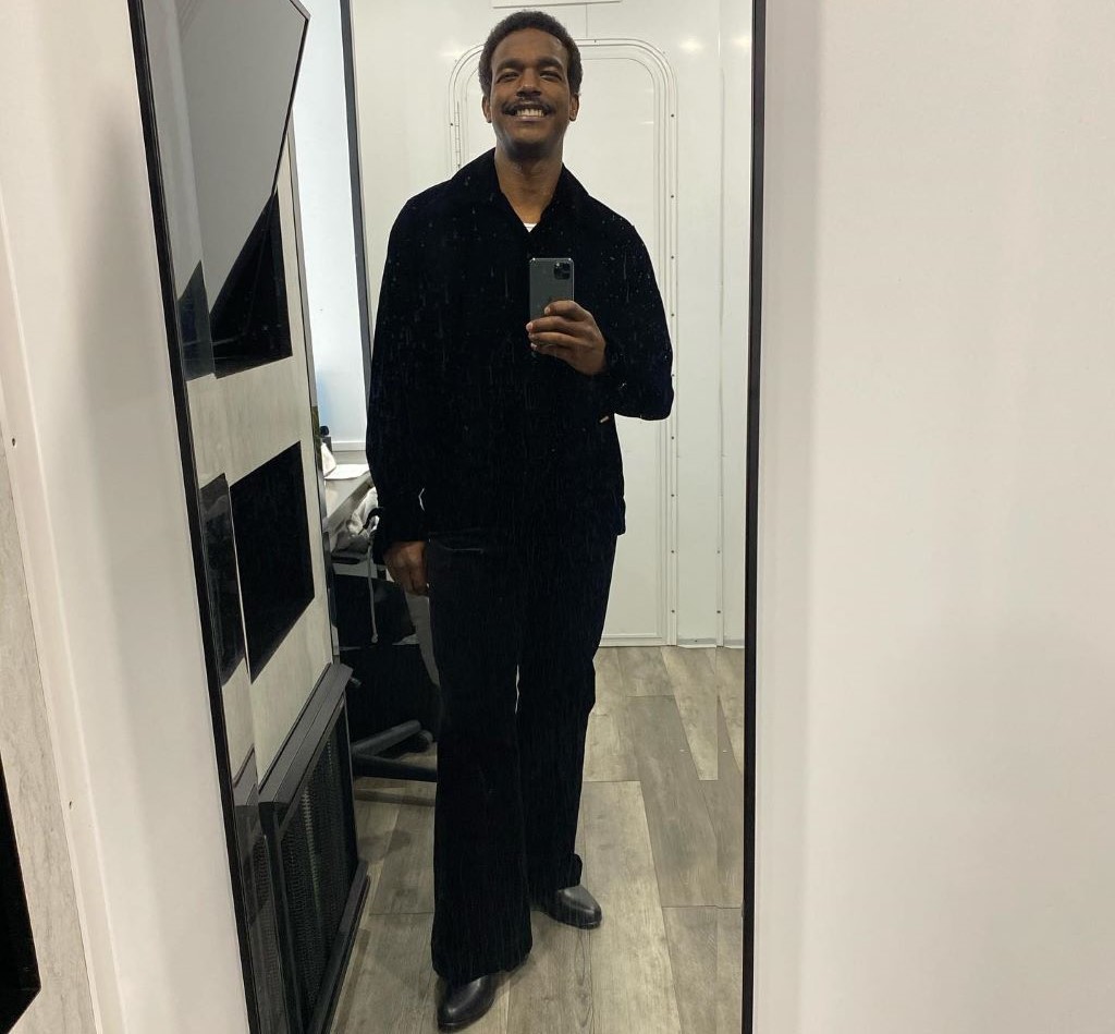 A mirror selfie of Luke James in which he is smiling. He is wearing full black colored outfit. 