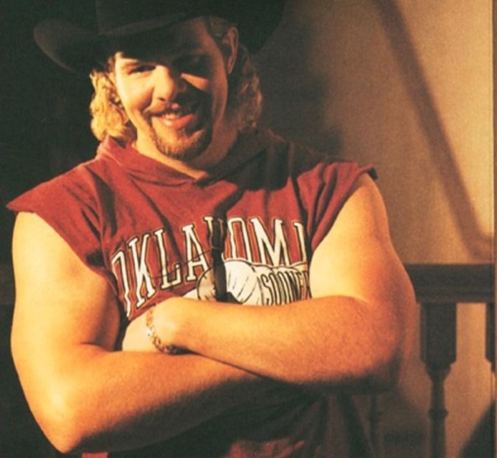 Toby Keith with no tattoo wearing sleeveless red shirt and hat smiling 