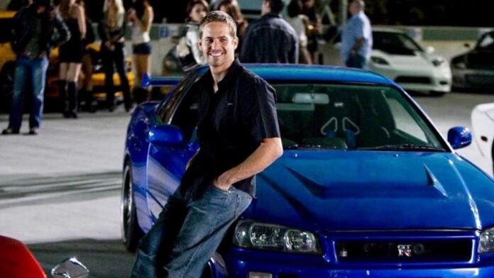 Paul Walker leans casually against a blue car, sporting a black dress, smiling, and striking a pose.