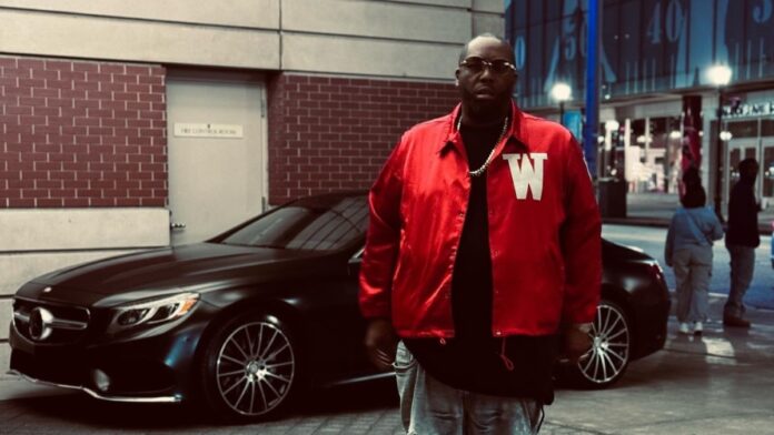 Killer Mike poses for a picture in front of a car wearing a red shirt, black vest, and goggles.