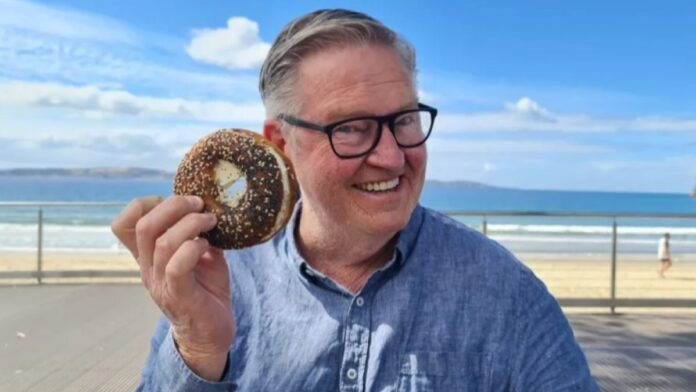 Peter poses with a sky-blue shirt, holding a chocolate doughnut in his hand.